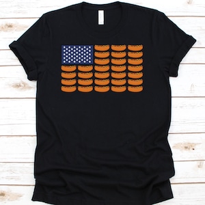 May include: Black t-shirt featuring a graphic of the American flag, with the stars replaced by white stars on a blue background and the stripes replaced by rows of hot dogs. The shirt has a crew neck and short sleeves.