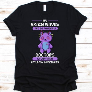 May include: Black t-shirt with a purple teddy bear graphic and text that reads "MY BRAIN WAVES ARE SO POWERFUL DOCTORS STUDY THEM EPILEPSY AWARENESS". The bear has button eyes and a heart on its chest.