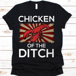 Puede incluir: Camiseta negra con un gráfico desgastado que presenta una langosta roja y las palabras "CHICKEN OF THE DITCH" en blanco. El diseño incluye un fondo de rayos de sol rojo y beige, creando un aspecto vintage.
