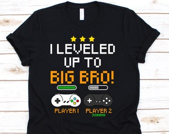 Puede incluir: Camiseta negra con el texto "I Leveled Up To Big Bro!" en fuente pixelada blanca y naranja. Debajo, dos gráficos de mandos de juego con las palabras "Player 1" y "Player 2 Joining".
