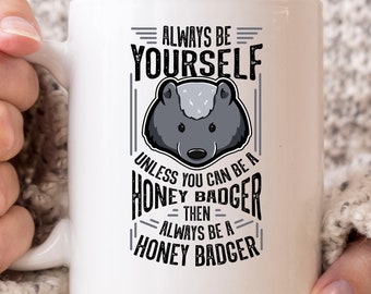 May include: White ceramic mug featuring a honey badger illustration and the text "ALWAYS BE YOURSELF UNLESS YOU CAN BE A HONEY BADGER THEN ALWAYS BE A HONEY BADGER". The mug has a white handle and is held by a person.