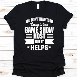 May include: Black t-shirt with white text. The text reads "YOU DON'T HAVE TO BE Crazy to be a GAME SHOW HOST but it HELPS". The design includes a banner and stars.
