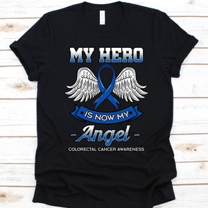 Puede incluir: Camiseta negra con el texto "MY HERO IS NOW MY Angel" en blanco y azul. En el centro se muestra una cinta azul y alas blancas. La parte inferior de la camiseta dice "COLORECTAL CANCER AWARENESS".