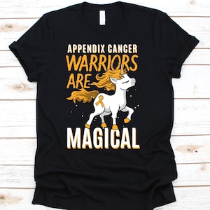 Puede incluir: Camiseta negra con el texto "APPENDIX CANCER WARRIORS ARE MAGICAL" en blanco y dorado. Se muestra un unicornio blanco con melena y cola doradas, junto con una cinta dorada.