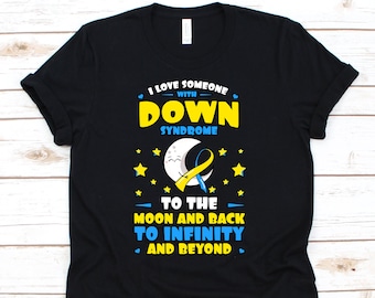 Op de afbeelding: Zwart t-shirt met de tekst "I LOVE SOMEONE WITH DOWN SYNDROME TO THE MOON AND BACK TO INFINITY AND BEYOND." Het ontwerp bevat een geel en blauw lint, sterren en een lachende maan.