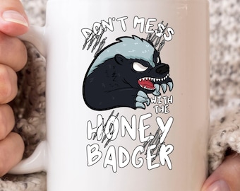 May include: White ceramic mug featuring a cartoon honey badger with an angry expression. The text "DON'T MESS WITH THE HONEY BADGER" is printed on the mug in a bold, playful font. The mug has a standard handle and is held by a person.