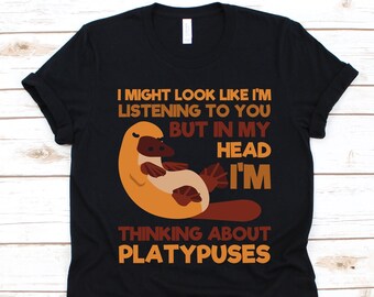 May include: Black t-shirt with a cartoon platypus illustration. The text reads, "I might look like I'm listening to you but in my head I'm thinking about platypuses." The design features orange, brown, and tan colors.