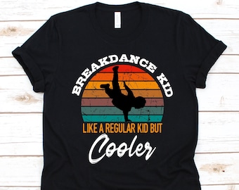 Puede incluir: Camiseta negra con una silueta de breakdance sobre un diseño retro de puesta de sol. El texto dice "BREAKDANCE KID" y "LIKE A REGULAR KID BUT Cooler" en blanco. Una camiseta informal para los fanáticos del baile.