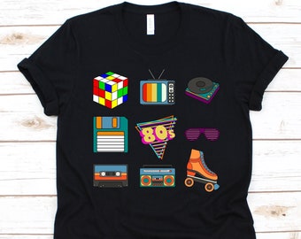 May include: Black t-shirt featuring colorful 80s-themed graphics. The design includes a Rubik's Cube, a retro TV, a record player, a floppy disk, the text "80s", sunglasses, a cassette tape, a boombox, and an orange roller skate.