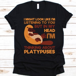 May include: Black t-shirt with a cartoon platypus illustration. The text reads, "I might look like I'm listening to you but in my head I'm thinking about platypuses." The design features orange, brown, and tan colors.