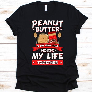 May include: Black t-shirt with a graphic featuring a peanut butter jar, a peanut butter sandwich, and the text "PEANUT BUTTER IS THE GLUE THAT HOLDS MY LIFE TOGETHER" in white and red. The design includes cartoon-style illustrations.