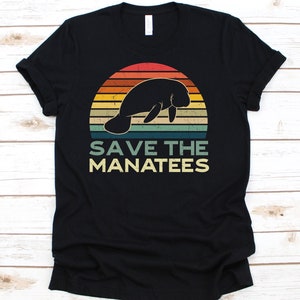 Puede incluir: Camiseta negra con un diseño retro. Presenta una silueta de manatí sobre un fondo de puesta de sol con rayas naranjas, amarillas y verdes. El texto "SAVE THE MANATEES" está impreso debajo en una fuente vintage.