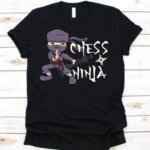 May include: Black t-shirt featuring a cartoon ninja with a chess piece and throwing star. The ninja is wearing a purple outfit and has the words "Chess Ninja" in white. The shirt is made of a soft material.