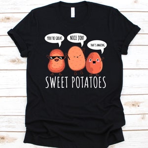以下が含まれることがあります： 黒いTシャツ。3つのサツマイモのイラストが描かれており、それぞれ腕と足があります。1つはサングラスをかけており、他の2つは「You're great」、「Nice job!」、「That's amazing」と書かれた吹き出しを持っています。「SWEET POTATOES」の文字が下に印刷されています。