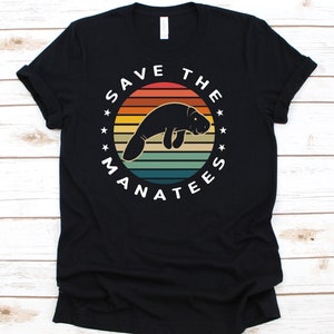 Puede incluir: Camiseta negra con una ilustración de manatí dentro de un diseño circular. El texto dice "SAVE THE MANATEES". El diseño incluye un fondo con colores de puesta de sol con rayas horizontales naranjas, amarillas y verdes.