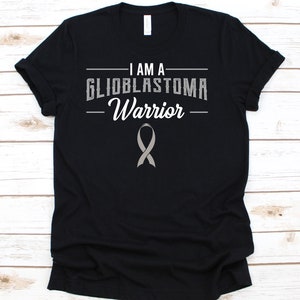 May include: Black t-shirt with the text "I AM A GLIOBLASTOMA Warrior" in white and grey. A grey ribbon is below the text. The shirt is made of a soft material.