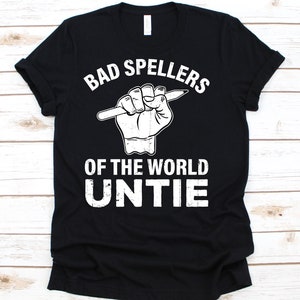 May include: Black t-shirt with the text "BAD SPELLERS OF THE WORLD UNTIE" in white. The graphic shows a hand holding a pencil. The shirt is made of a soft material and has a classic crew neck.