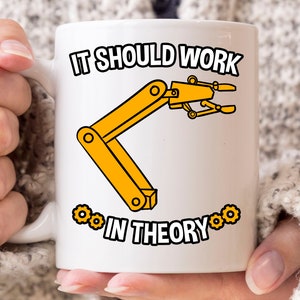 Op de afbeelding: Witte keramische mok met een gele robotarm illustratie en de tekst "IT SHOULD WORK IN THEORY". Het ontwerp bevat gele tandwielen. De mok wordt door een persoon vastgehouden.