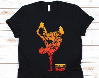 Puede incluir: Camiseta negra con una silueta de breakdancer en tonos naranja y amarillo, compuesta por varios objetos. Un gráfico de radio retro en la parte inferior. La camiseta tiene cuello redondo.