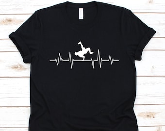Puede incluir: Camiseta negra con un gráfico blanco de un breakdancer incorporado en una línea de latido cardíaco. La camiseta está hecha de un material suave y tiene un cuello redondo clásico.