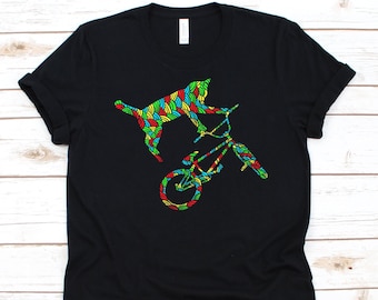 May include: Black t-shirt featuring a colorful graphic of a cat riding a BMX bike. The cat and bike are filled with a vibrant, patterned design in shades of red, yellow, green, and blue. The shirt is a classic crew neck style.