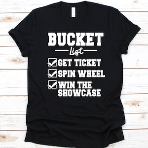 Può includere: T-shirt nera con testo bianco che recita "BUCKET List" sopra un elenco. L'elenco include "GET TICKET", "SPIN WHEEL" e "WIN THE SHOWCASE", ognuno con un segno di spunta.
