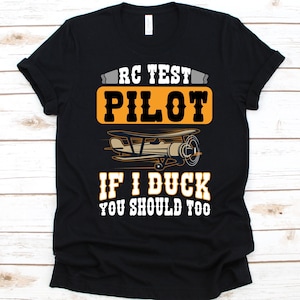 May include: Black t-shirt with a graphic design featuring the text "RC TEST PILOT" above an illustration of a biplane. Below the plane, the text reads "IF I DUCK YOU SHOULD TOO". The design is in white, orange, and tan.