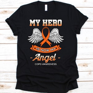 Puede incluir: Camiseta negra con el texto "MY HERO is now my Angel" en blanco y naranja. El diseño incluye una cinta naranja con alas blancas y las palabras "COPD AWARENESS".
