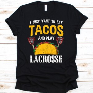 May include: Black t-shirt with the text "I JUST WANT TO EAT TACOS AND PLAY LACROSSE" in yellow and white. A graphic of a taco and lacrosse sticks is also featured on the shirt.