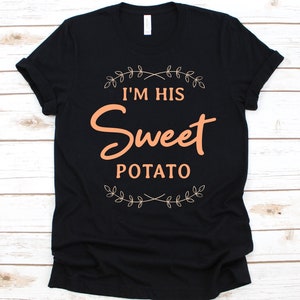 以下が含まれることがあります： 黒いTシャツ。桃色の文字で「I'M HIS Sweet POTATO」と書かれており、葉の装飾が施されています。柔らかい素材で作られたカジュアルウェアです。