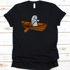 May include: Black t-shirt with a cartoon manatee rowing a wooden boat. The manatee is grey, and the boat is brown. The design is centered on the shirt, offering a playful nautical theme.