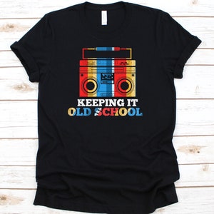 May include: Black t-shirt featuring a retro boombox graphic in red, yellow, blue, and white. The text "KEEPING IT OLD SCHOOL" is below the boombox. A classic design for music lovers.