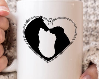 Könnte beinhalten: Weiße Keramiktasse mit einer schwarzen Silhouette eines Pferdes und eines Hundes in einem herzförmigen Design. Das Design hat einen dekorativen Rand und ist auf der Tasse zentriert. Die Tasse hat einen gebogenen Griff und wird von einer Person gehalten.