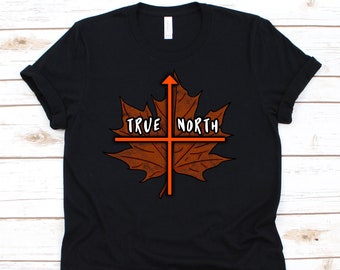 Puede incluir: Camiseta negra con un diseño de hoja de arce marrón, una brújula naranja y las palabras "TRUE NORTH" en blanco. La camiseta tiene mangas cortas y cuello redondo.
