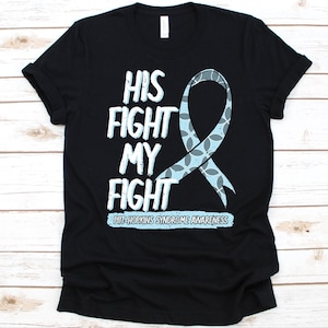 Puede incluir: Camiseta negra con el texto "HIS FIGHT MY FIGHT" en blanco y azul claro. Se presenta una cinta azul claro con un patrón. El texto "PITT-HOPKINS SYNDROME AWARENESS" está debajo.