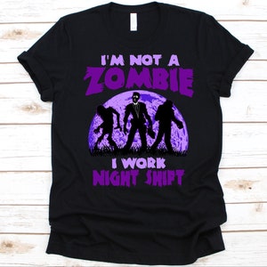 May include: Black t-shirt with a purple graphic that reads "I'M NOT A ZOMBIE I WORK NIGHT SHIFT." The graphic features three zombie silhouettes in front of a purple moon.