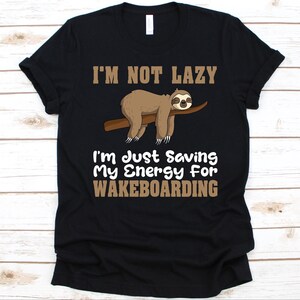 May include: Black t-shirt with a brown sloth graphic on a branch. The text reads "I'M NOT LAZY I'm Just Saving My Energy For WAKEBOARDING". The shirt is made of a soft material.