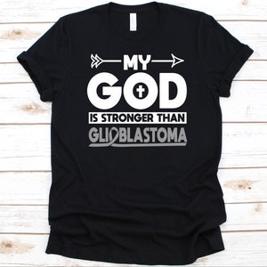 May include: Black t-shirt with white text that reads "MY GOD is STRONGER THAN GLIOBLASTOMA." The word "GOD" has a cross in the center. The text is aligned with an arrow pointing to the right.