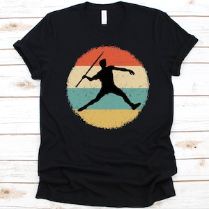 May include: Black t-shirt featuring a retro design of a javelin thrower in silhouette. The design is set within a distressed circle with horizontal bands of red, teal, and yellow. A classic athletic-themed tee.