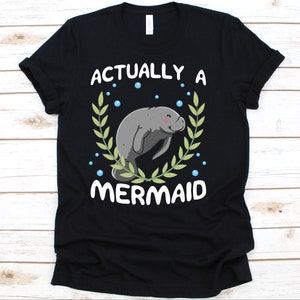 May include: Black t-shirt with the text "ACTUALLY A MERMAID" in white. The graphic features a grey manatee with a smiling face, surrounded by green leaves and blue bubbles.