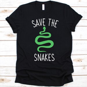 May include: Black t-shirt with the text "SAVE THE SNAKES" in white, above a green snake illustration. The snake is a stylized, curved design. The shirt is a crew neck style.
