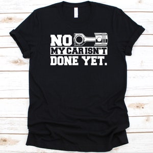 May include: Black t-shirt with white text and a piston graphic. The text reads "NO, MY CAR ISN'T DONE YET." The shirt is a crew neck style.