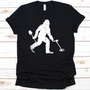 Könnte beinhalten: Schwarzes T-Shirt mit einem weißen, verwitterten Aufdruck von Bigfoot beim Metalldetektieren. Bigfoot hält eine Schaufel und einen Metalldetektor. Das T-Shirt ist aus einem weichen Material.