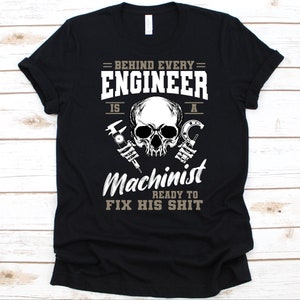 May include: Black t-shirt with a white and grey graphic. The graphic features a skull, calipers, and a wrench, with the text "BEHIND EVERY ENGINEER IS A MACHINIST READY TO FIX HIS SHIT."