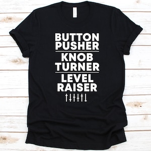May include: Black t-shirt with white text: "BUTTON PUSHER, KNOB TURNER, LEVEL RAISER" and graphic of sliders. The shirt is made of a soft material and has a crew neck.