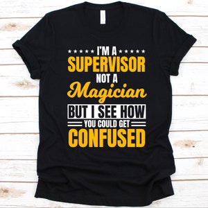 以下が含まれることがあります： 黒いTシャツ。黄色と白で「I'M A SUPERVISOR not a Magician BUT I SEE HOW YOU COULD GET CONFUSED」というテキストが書かれています。テキストは積み重ねられた形式で配置されています。