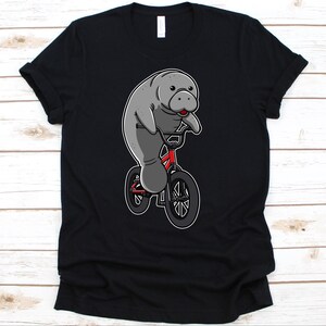 May include: Black t-shirt with a cartoon manatee riding a red bicycle. The manatee is grey with a red tongue, and the bicycle has black tyres. The design is centered on the shirt.