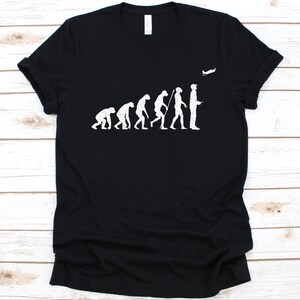 Może przedstawiać: Czarny t-shirt z białym nadrukiem przedstawiającym ewolucję człowieka, zakończoną osobą obserwującą mały samolot. Projekt oferuje humorystyczne spojrzenie na rozwój lotnictwa.