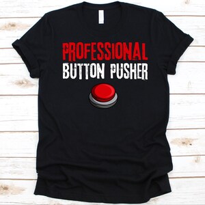 May include: Black t-shirt with the text "PROFESSIONAL BUTTON PUSHER" in red and white. A red button graphic is below the text. The shirt is made of a soft material.