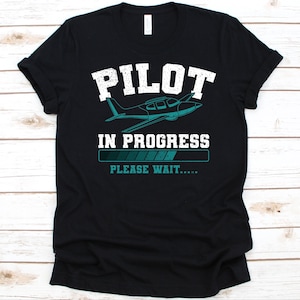 May include: Black t-shirt with the text "PILOT IN PROGRESS" above an airplane graphic and a loading bar. Below the loading bar is the text "PLEASE WAIT.....". The text and airplane are in white and teal.
